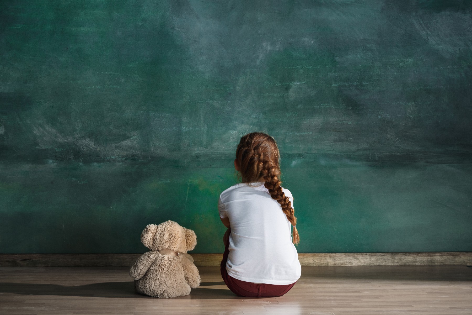 Little girl with teddy bear sitting on floor in empty room. Autism concept