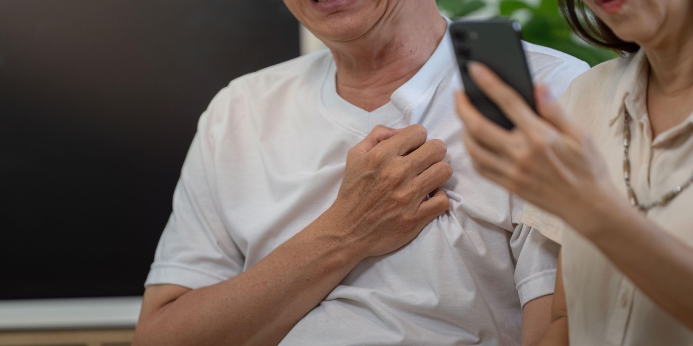 Elderly man smiling while holding his chest, woman on phone in supportive role.