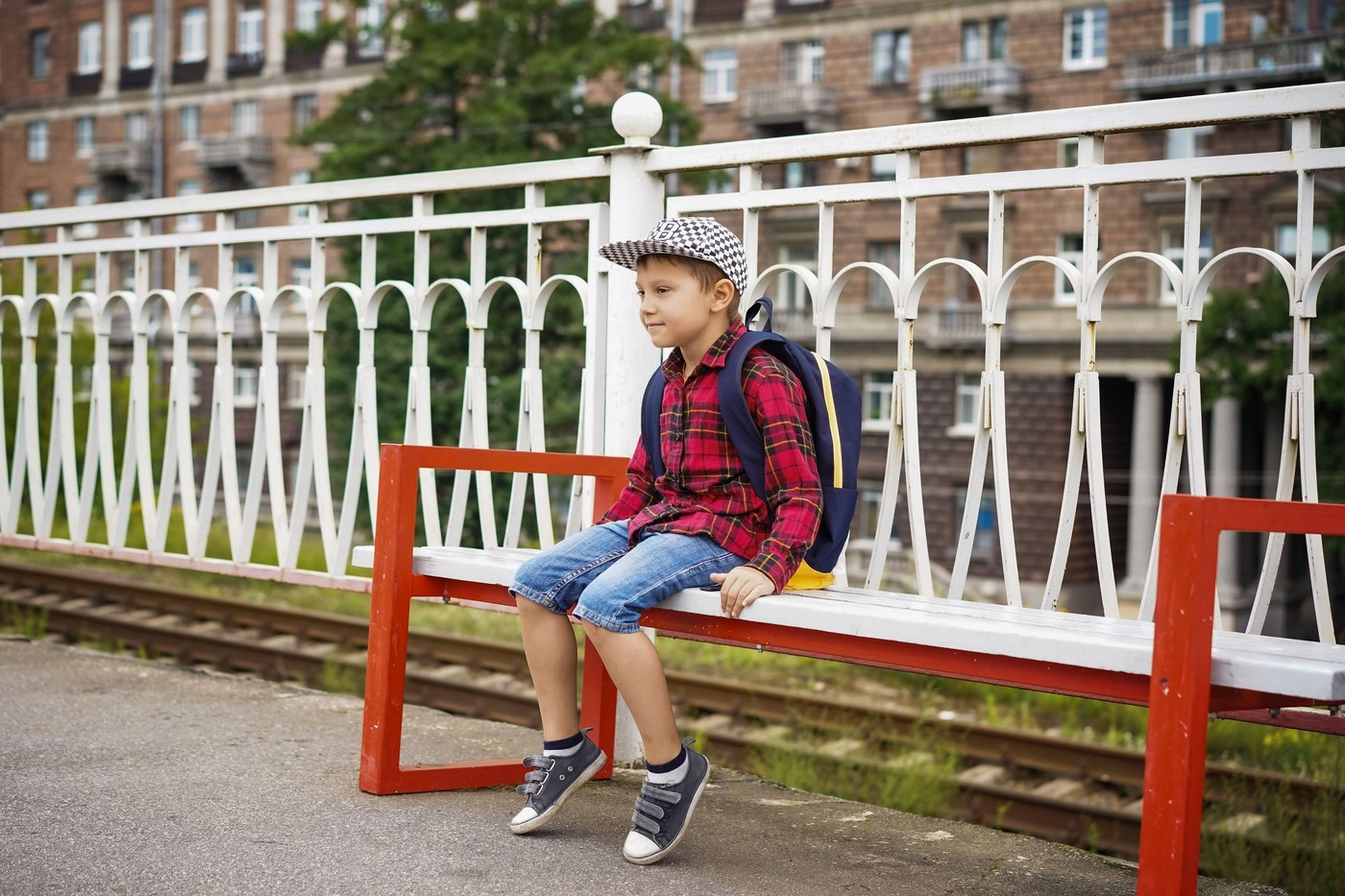 cute caucasian boy in checkered shirt, cap and jean shorts sitting on bench at rail station waiting for the train with pensive face. High quality phot