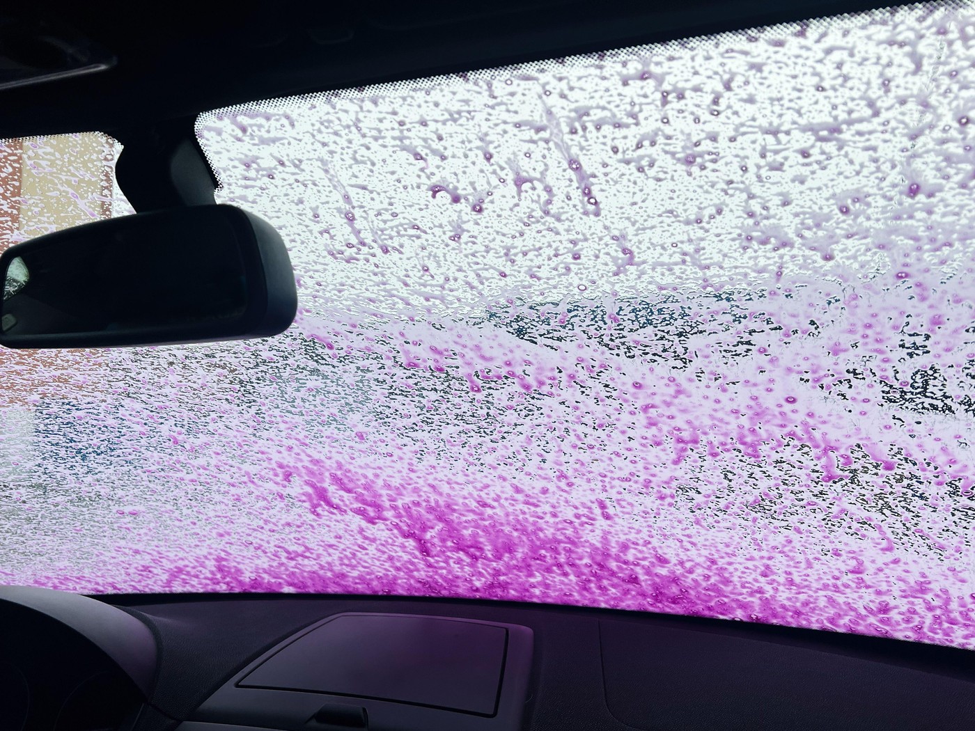 Foam-covered windshield of a car viewed from the interior during a lively car wash, showcasing vibrant pink and white soap bubbles.