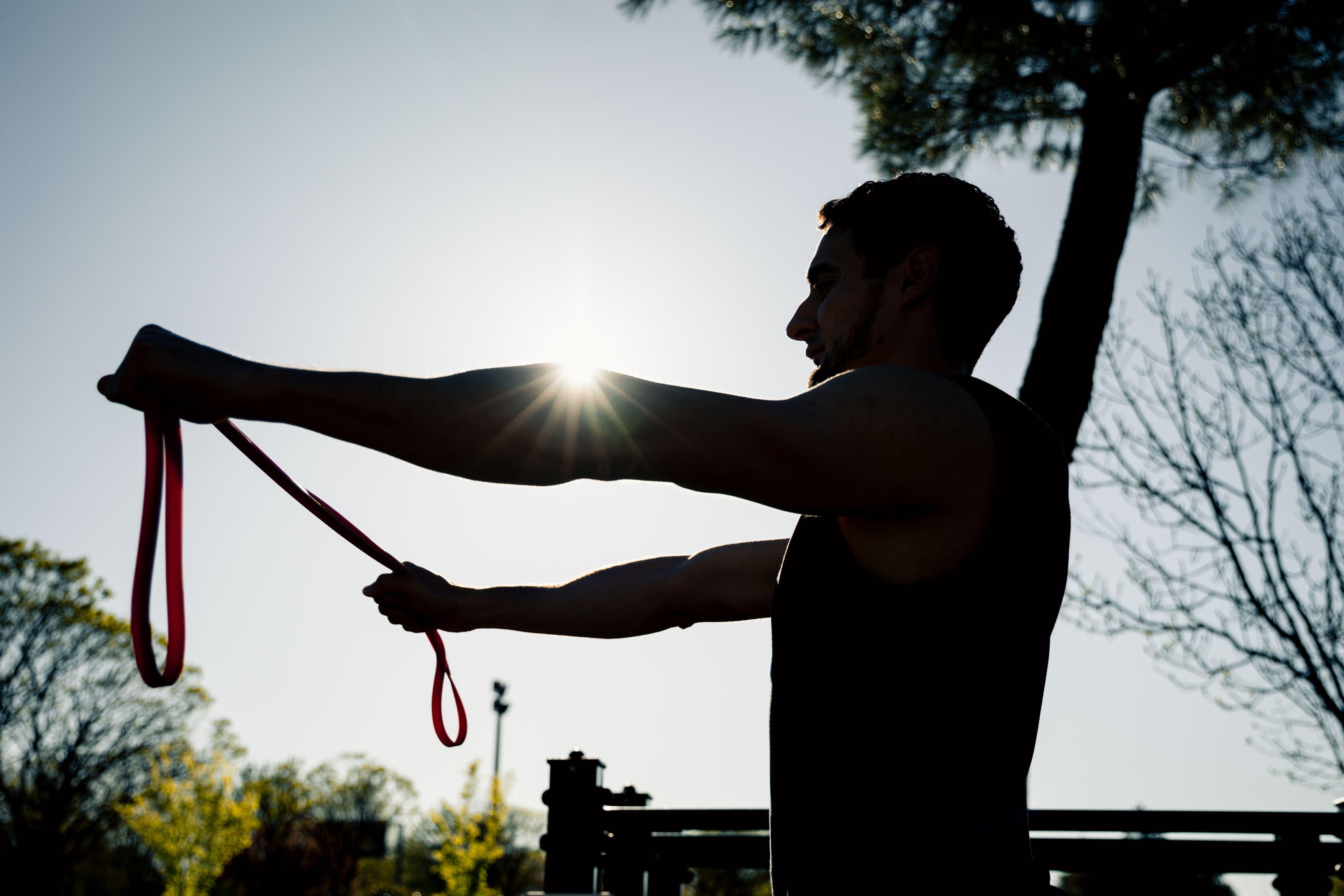Silhouette of athletic man exercising with red resistance band outdoors at sunrise, demonstrating strength, focus, and workout dedication in a natural