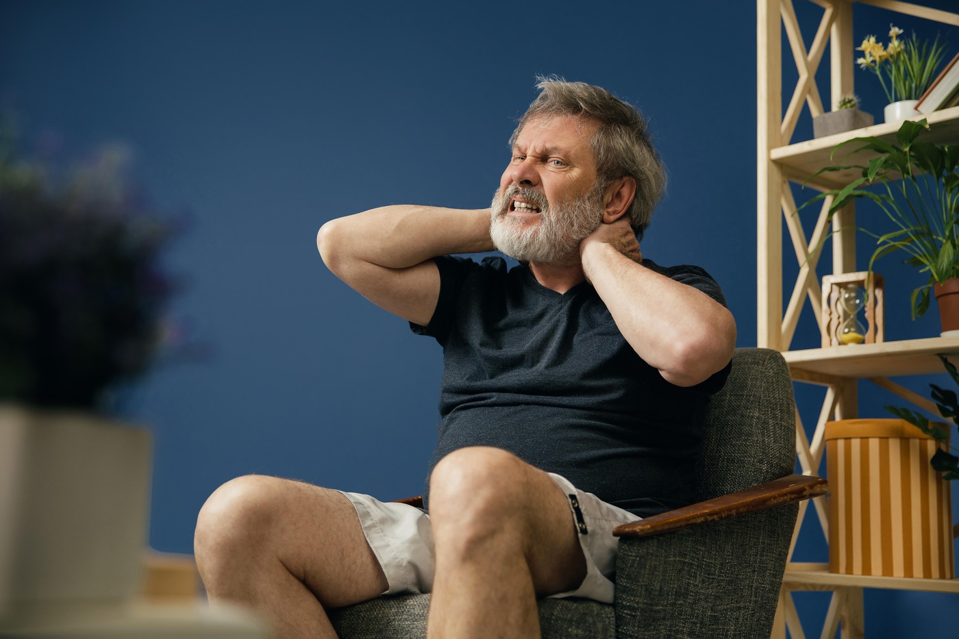 Life's getting hard. Old bearded man sitting on the chair and suffering from neck pain on blue background. Concept of illness, diseases of the joints and bones, surgical pathology, healthcare.