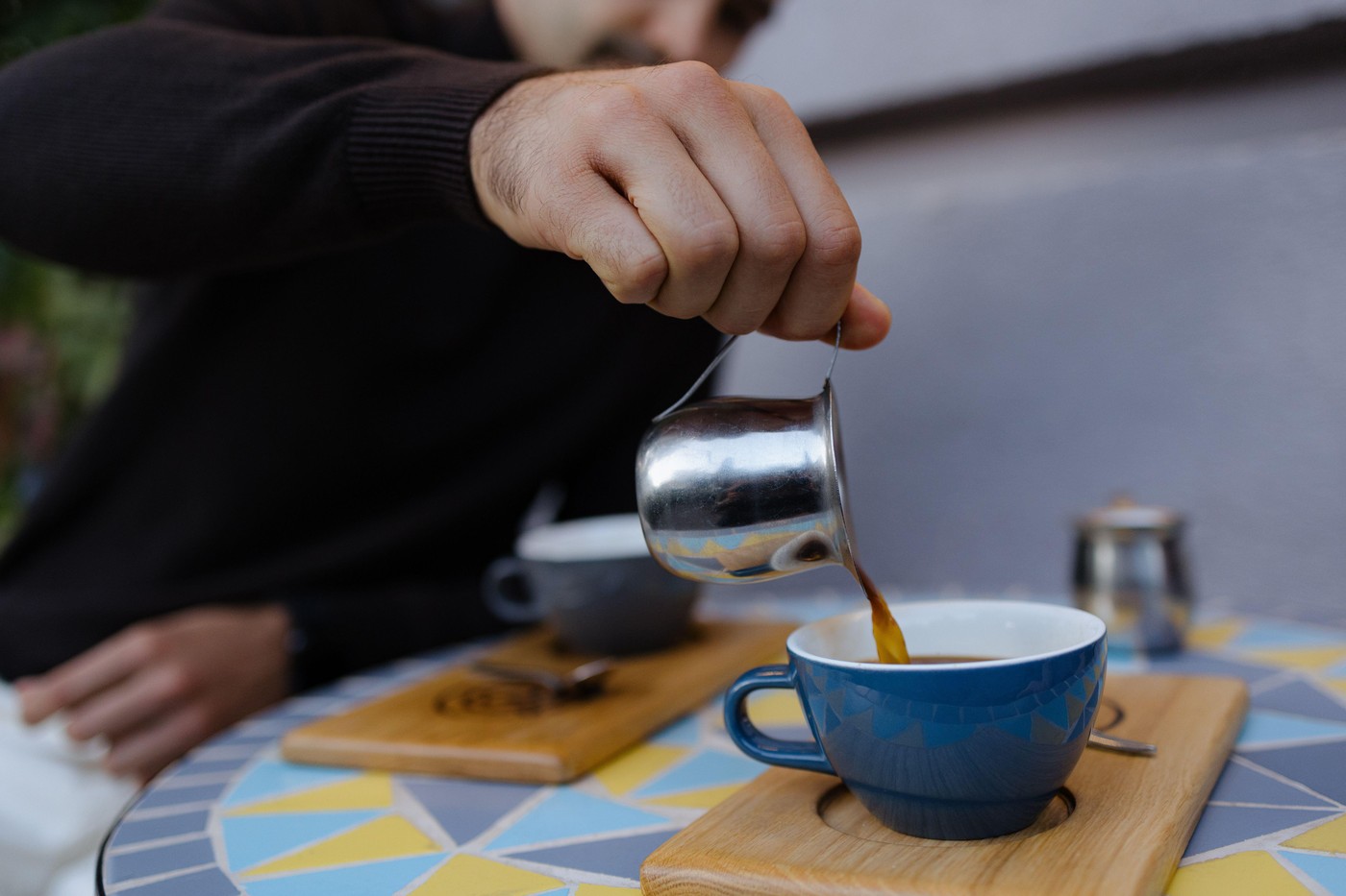 A person elegantly pours steaming coffee into a charming blue cup on a colorful tiled table. The atmosphere is warm and inviting, perfect for a relaxe