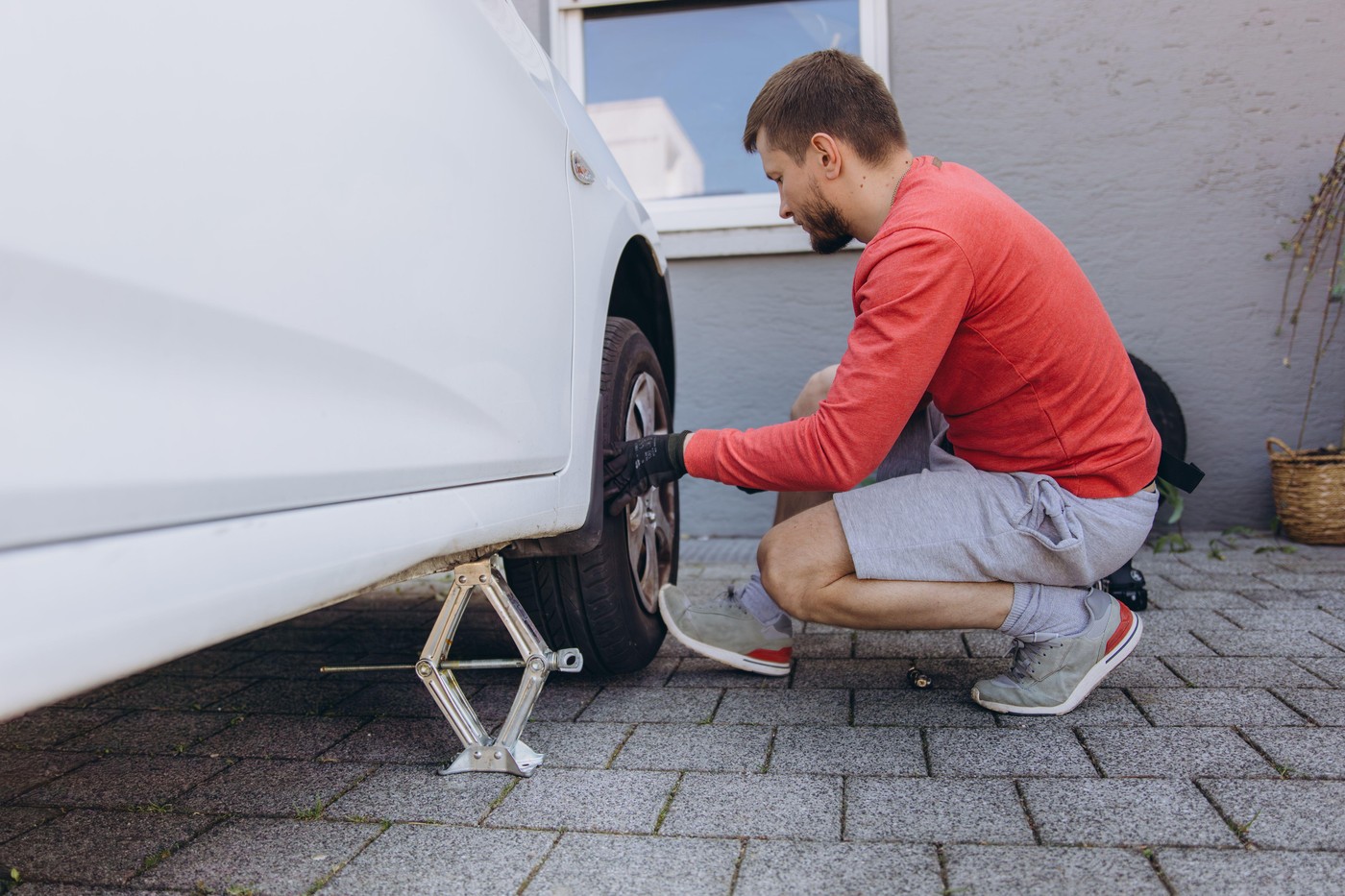 Close-up of a man changing a car tire in front of his house. The vehicle is lifted with a jack, tools lie nearby. The scene captures a hands-on repair