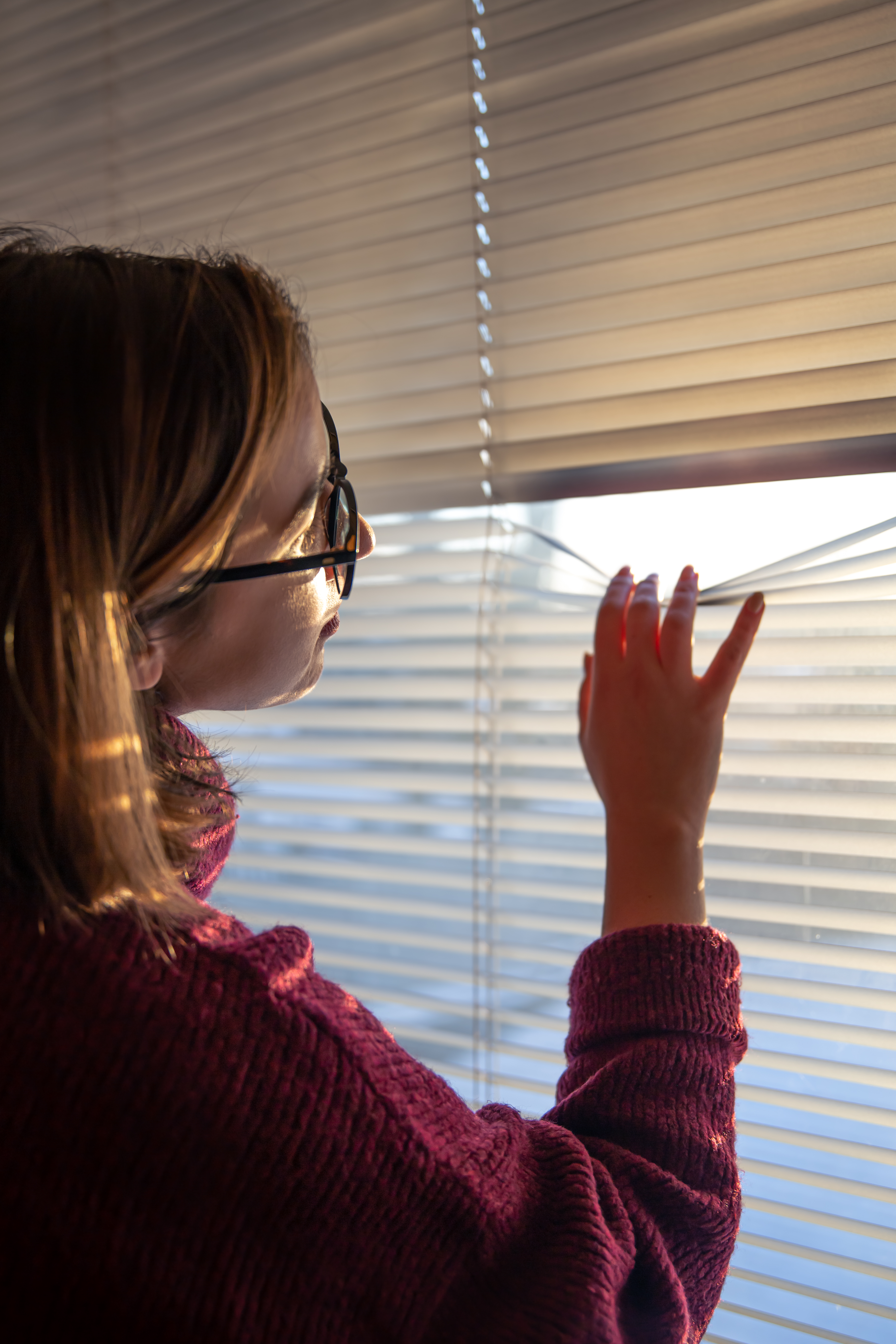 A woman with glasses looks through the blinds at the early morning sunlight.