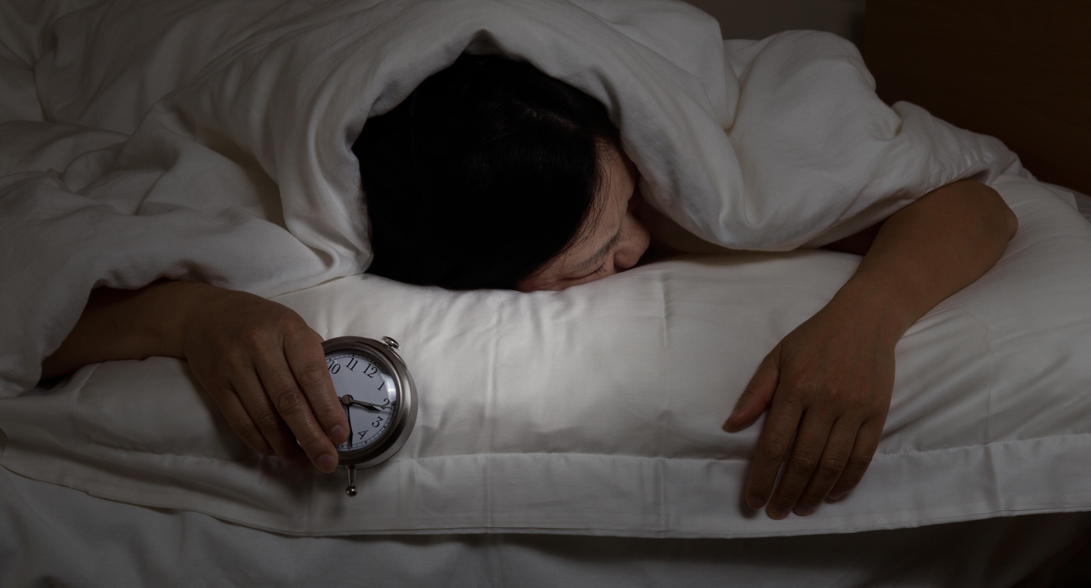 Woman with insomnia, under blanket looking down, holding alarm clock in hand.